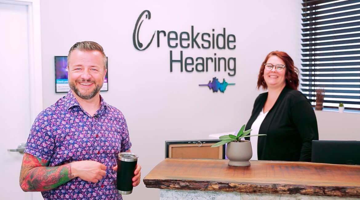 Patient with Michelle, owner of Creekside Hearing, laughing at front desk with a coffee in his hand