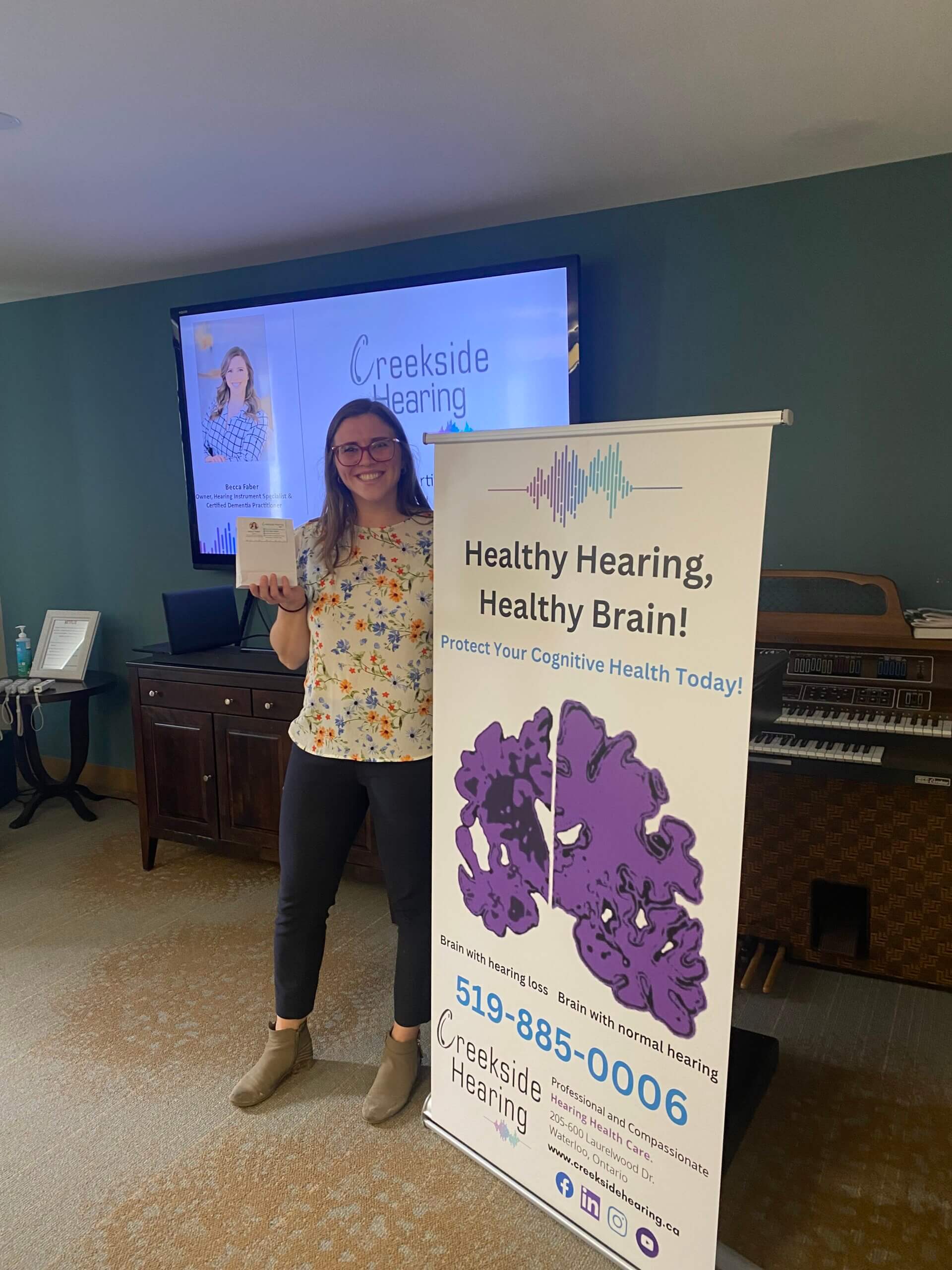 Becca from Creekside Hearing, standing in front of a banner with a photo of a brain with hearing loss at Court at Laurelwood